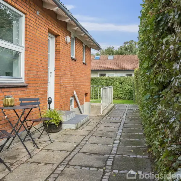 En terrasse med bord og stole i træ ved siden af en rød murstensbygning. Baggrunden har en grøn hæk, og en flisebelagt sti fører til en trappe ved døren.
