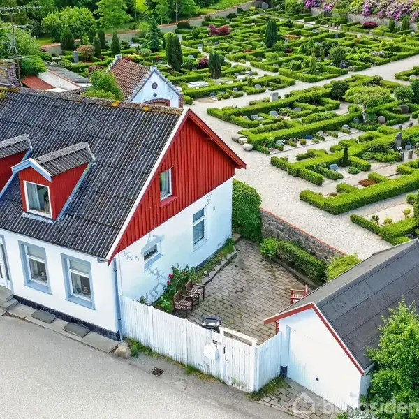 Hvidt hus med røde vinduer og døre, stående ved en vej med brosten. En velplejet have med symmetriske buske ligger bagved, omgivet af grønne træer og blomster.