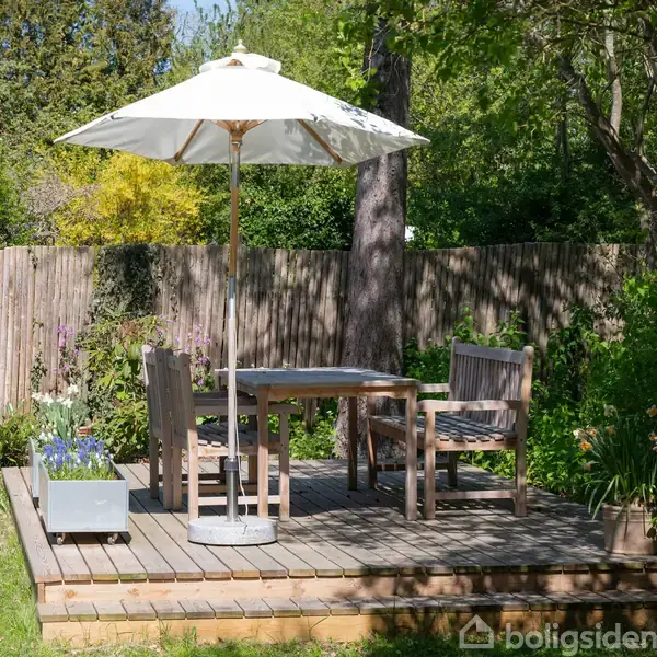 En træterrasse med et hvidt parasol, et bord og fire stole. Omkring terrassen er en frodig have med buske og træer, indrammet af et træhegn.