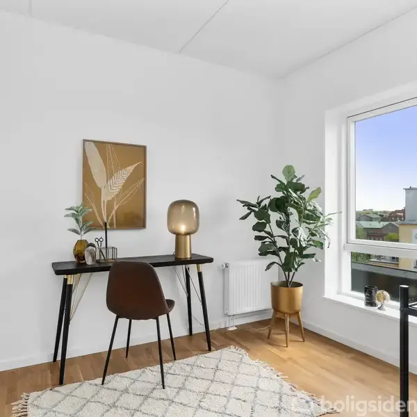 Skrivebord med stol står i et lyst rum. En lampe og planter pynter bordet. Vinduet viser udsigt til bygninger og blå himmel udenfor. En tæppe ligger på trægulvet.