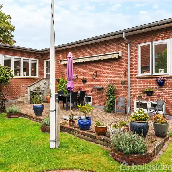 Rød murstensvilla med terrasse, hvor der står et bord med stole og en parasol. Haven er grøn og velplejet med forskellige planter og blomsterkrukker.