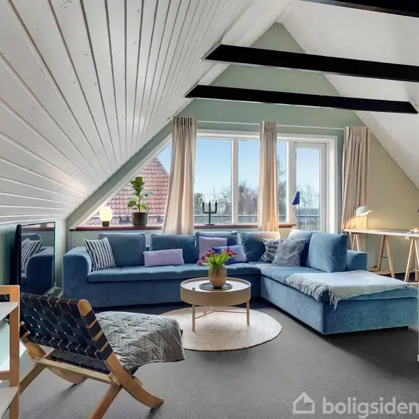 Stue med blå hjørnesofa under skråloft. Sofabord med blomster foran, omgivet af puder. Store vinduer i baggrunden med udsigt, hvid vægreol til venstre og lænestol med tæppe.