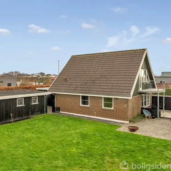 Et murstenshus med trekantet tag står i en have med grønt græs. Ved siden af er en sort træskur samt en terrasse med møbler. Himlen er blå med få skyer.