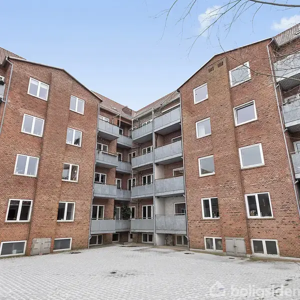 En murstensejendom med flere etager, hvor hver lejlighed har balkon. Bygningen omkranser en brostensbelagt gårdsplads, mens træer og himmel ses i baggrunden. Ingen tekst er til stede.