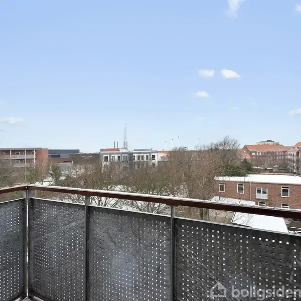 En balkon med perforeret metalrækværk står stille, omgivet af en byudsigt med flade bygninger og træer under en let skyet himmel.