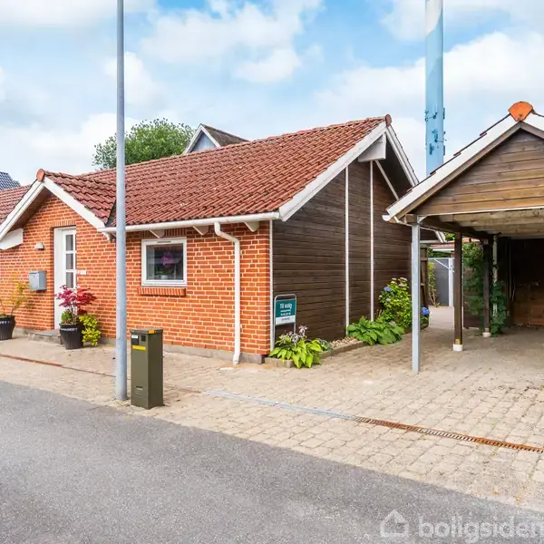 Rødt murstenshus med rødt tag står ved siden af et åbent carport i et forstadskvarter. Huset har hvidrammede vinduer og en blomsterpotte ved indgangen.