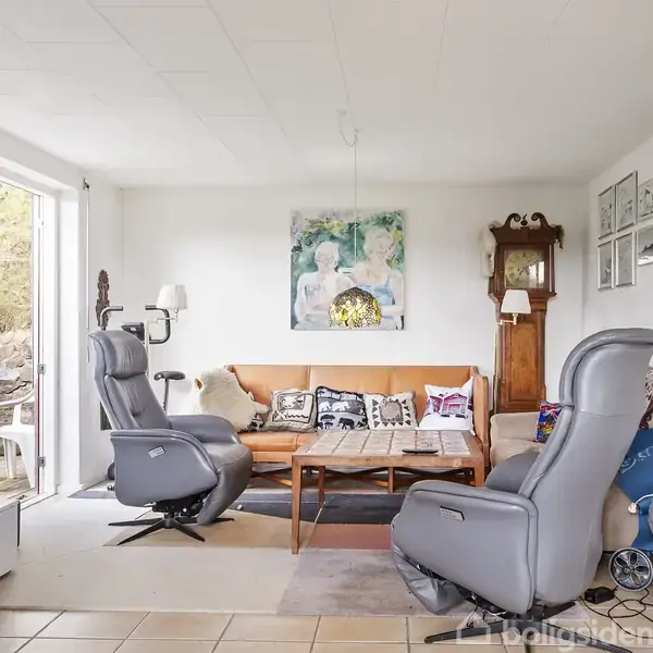 To lænestole vender mod en sofa med farverige puder, stående foran et sofabord. Væggen har malerier og fotografier, og en stor glasdør åbner mod en terrasse.