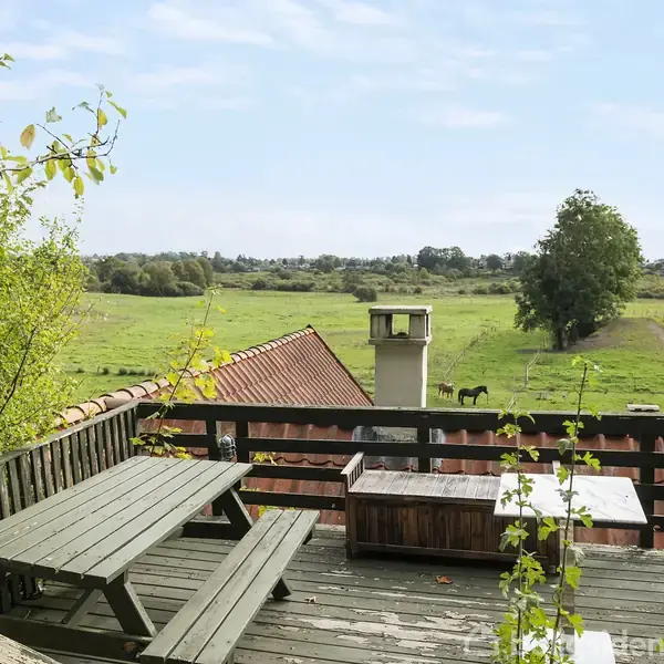 En træterrasse med et picnicbord og bænk står udenfor. I baggrunden ses en grøn mark med enkelte træer og en hest, under en blå himmel med spredte skyer.