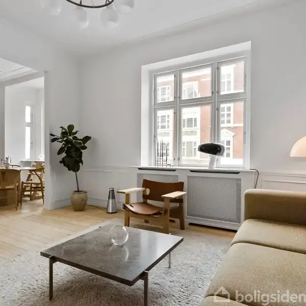 En lys stue med beige sofa, sofabord og gulvlampe. Et stort vindue giver udsigt til en bygning udenfor. I baggrunden ses et spisebord og stole på trægulv.