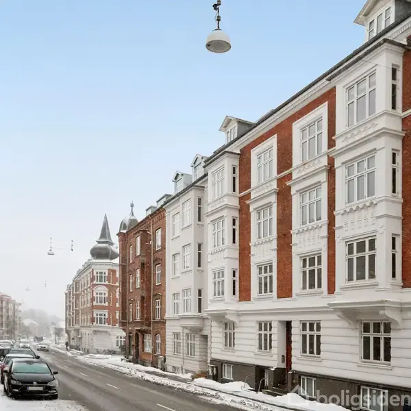 Bygninger med røde og hvide facader står langs en snedækket gade med parkerede biler og gadelygter i en urban vinterkontekst.