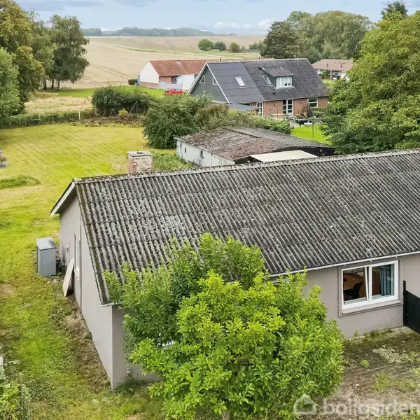 Et enetages hus med gråt tag står i en grøn have omgivet af buske. I baggrunden ses flere huse og åbne marker under en skyet himmel.