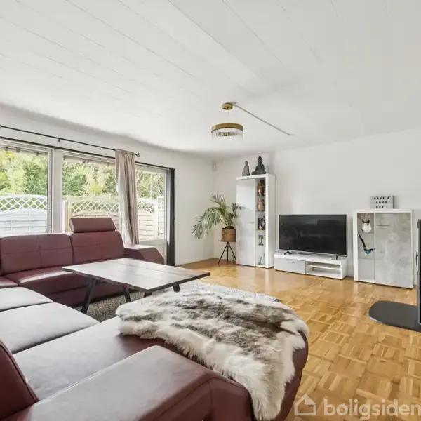 En stue med en rødbrun hjørnesofa dækket af et tæppe. Foran sofaen står et sofabord. Ved siden af er en brændeovn og et tv på en skænk. Store vinduer slipper lys ind.