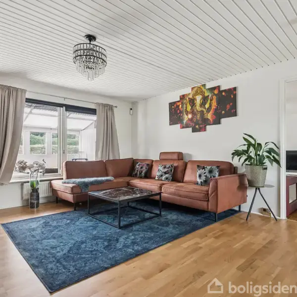 Brunt hjørnesofa står på et blåt tæppe i en stue, med et sofabord foran. Vinduer med gardiner giver udsigt til haven. En moderne lysekrone hænger fra loftet.