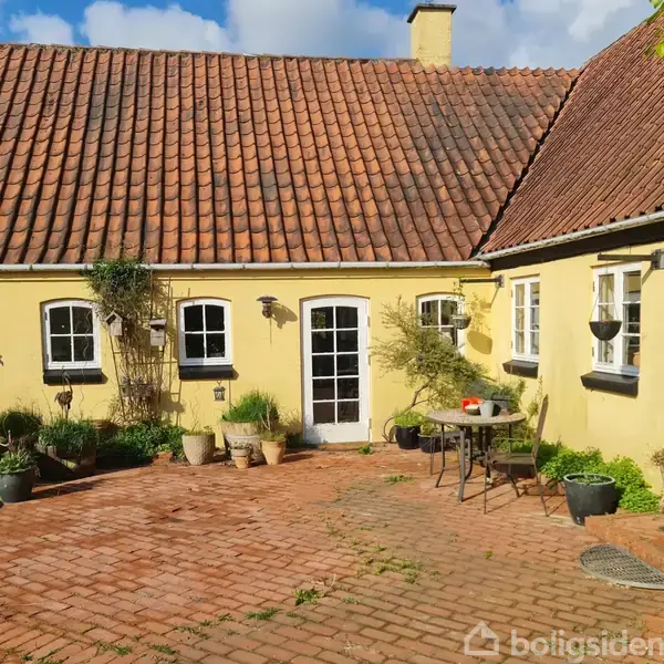 Gult murstenshus med rødt tegltag omkranser en brostensbelagt gårdsplads. Potteplanter er placeret rundt om gården, og et lille bord og stole står tæt på den centrale glasdør.