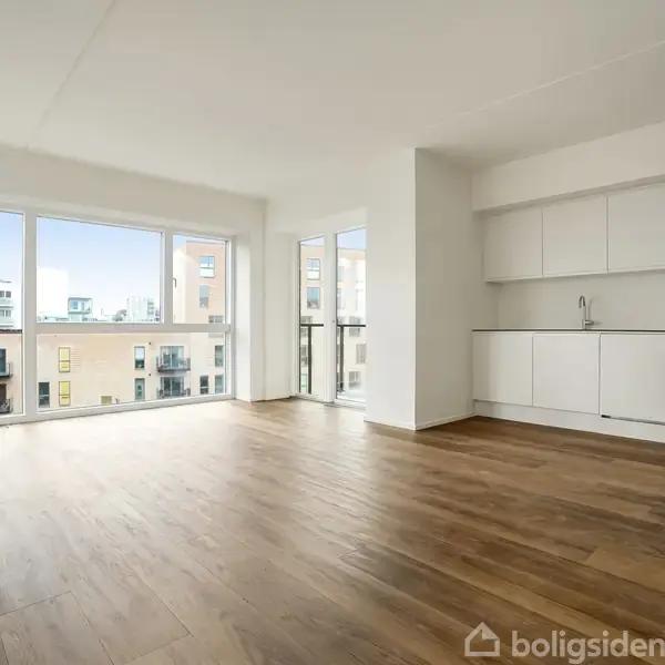 Lejlighedsstue med trægulv og store vinduer, der ud kigger over nærliggende bygninger. Moderne, hvidt køkken uden apparater står langs væggen, skaber en rummelig og lys boligindretning.