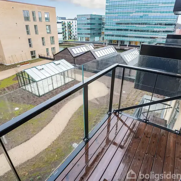 En altan med glasrækværk i en moderne bygning vender ud mod en gård med stier og drivhuse. I baggrunden ses flere kontor- og boligkomplekser.