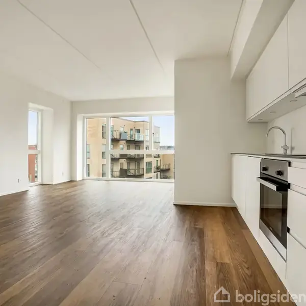 Et moderne køkken med hvide skabe og indbygget ovn støder op til en rummelig stue med trægulve. Store vinduer viser udsigt til balkon og nabobygninger.