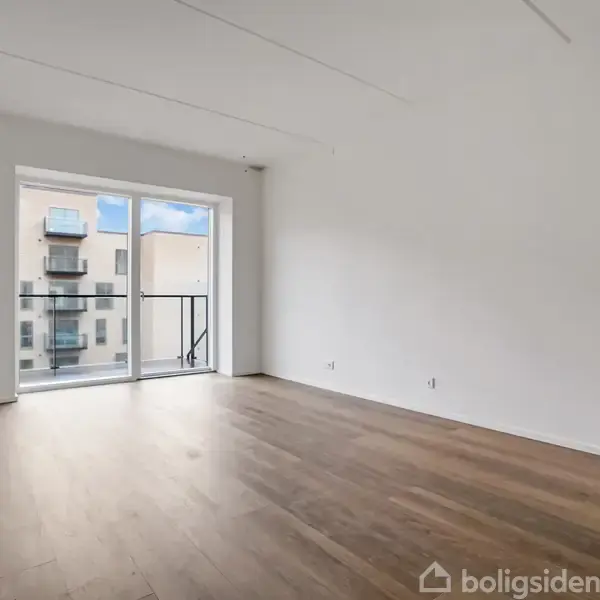 Et tomt værelse med træparketgulv, hvor sollys skinner ind gennem en stor skydedør til en lille altan. Udenfor ses en moderne bygning. Ingen tekst er til stede.