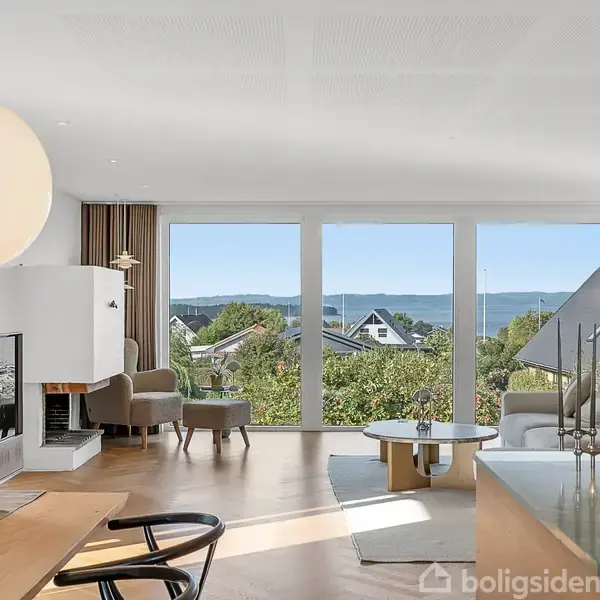 En lys og rummelig stue med moderne møblering, hvor sollys strømmer ind gennem store vinduer med udsigt over havet og huse i baggrunden. En pejs og blomsterdekoration pryder rummet.