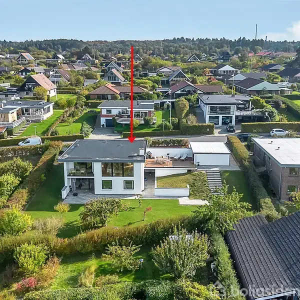 Et hvidt, moderne hus med fladt tag og store vinduer ligger i frodige omgivelser. Rød pil peger ned på huset, omgivet af flere villaer og grøn have.