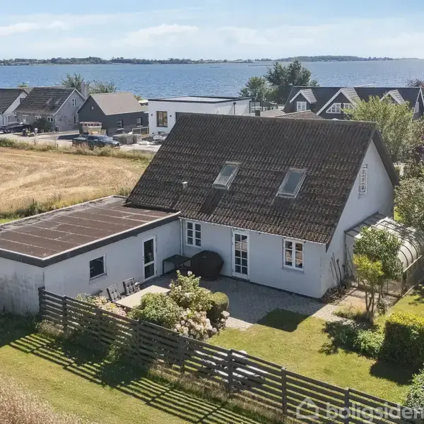 Et hvidt hus med skråt tag står stille omgivet af en velplejet have, en lav træhegn og en terrasse. I baggrunden ses en kystlinje med flere huse.