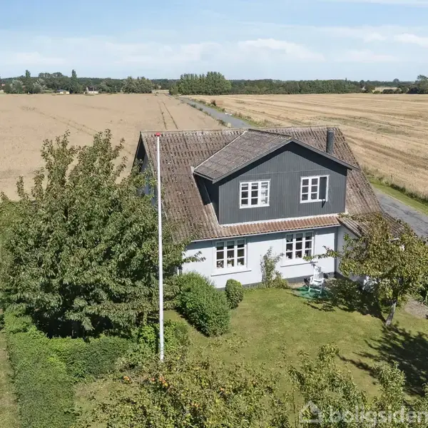Et to-etagers hus omgivet af træer står i en stor mark med en vej til højre. Husets facade er hvid med gråt gavlparti og traditionelle vinduer. Markerne strækker sig langt ud.