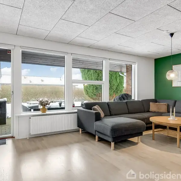 En stor, grå hjørnesofa står i en stue op ad en grøn væg med en rund træbord foran. Rummet har store vinduer, som giver udsigt til en have med en terrasse.