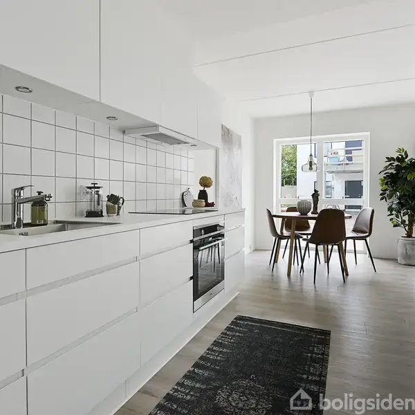 Et lyst køkken med hvide skabe og fliser, hvor der står planter og køkkenredskaber. I baggrunden ses et spisebord med stole ved et vindue og en stor plante ved siden af.