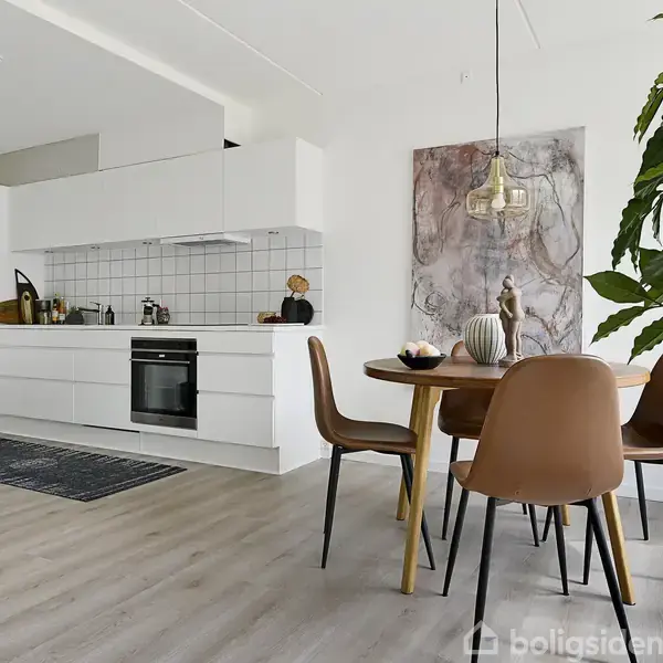 Et moderne køkken med hvide skabe og integreret ovn. Ved siden af står et rundt spisebord med fire brune stole. En stor plante til højre, og på væggen hænger et maleri.