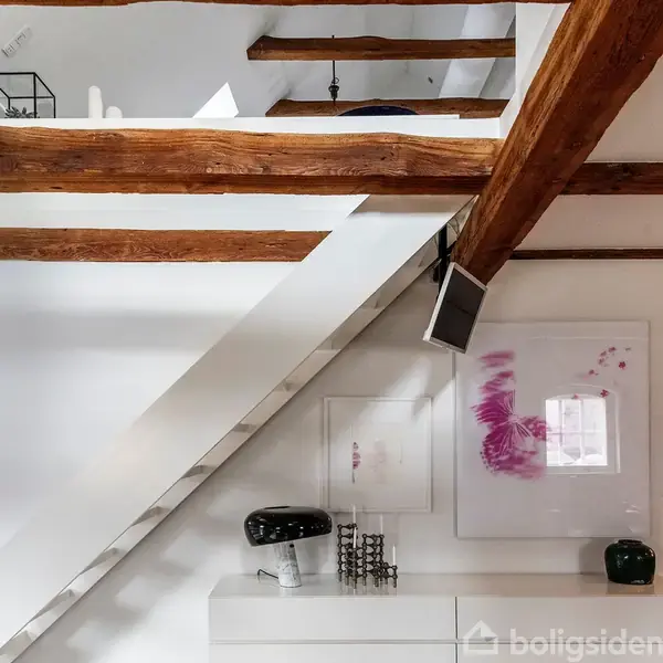 Bjælker i loftet kombineret med moderne hvide vægge. En TV-skærm hænger skråt under trappen. Dekorative billeder med pink-motiv pryder væggen, og en sort lampe står nedenfor.