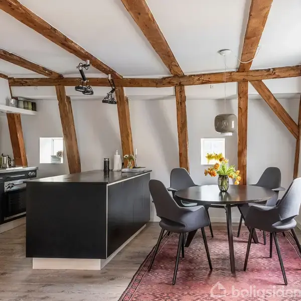 Et moderne køkken med synlige træbjælker har en sort køkkenø og et rundt spisebord med fire stole. Rummet er lyst med hvide vægge og et trægulv.