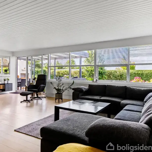 Stor hjørnesofa i mørkegråt står i en stue med lyse trægulve. Store vinduer giver udsigt til en grøn have. Et sofabord står foran sofaen, og der er en fjernsynsskærm til venstre.