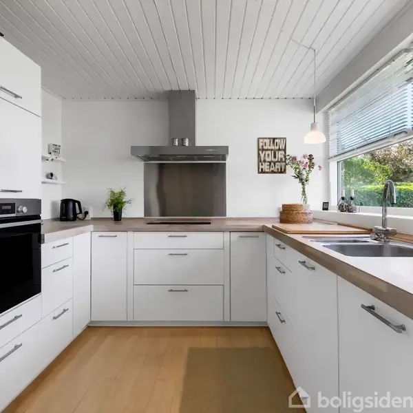 Moderne køkken med hvide skabe og indbygget ovn. Grå emhætte over komfuret. Tekst på væg: "FOLLOW YOUR HEART". Lysindfald gennem store vinduer. Blomsterdekoration og elkedel på bordet.