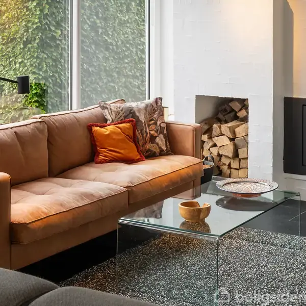 Brun sofa står i en stue med glasbord foran. Ved siden af er en pejs med ild, og brænde er stablet i en niche. Store vinduer viser en grøn plantevæg udenfor.