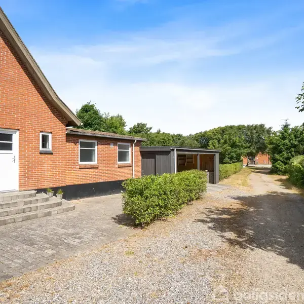 Rødt murstenshus med hvide vinduer står stille ved siden af en grusvej. Foran huset er en flisebelagt indgang og en lille hæk. Vegetation omgiver området.