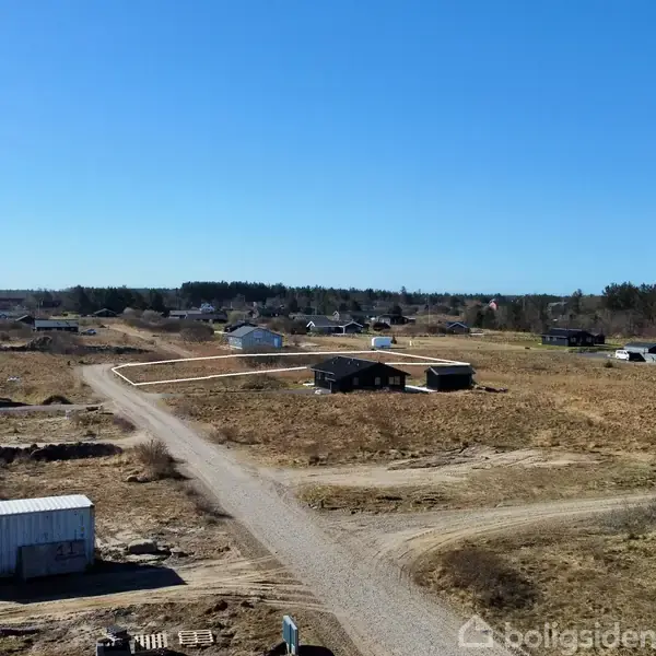 Landområde med opførte huse og en grusvej, omgivet af åbne marker og skov i baggrunden under en klar blå himmel.