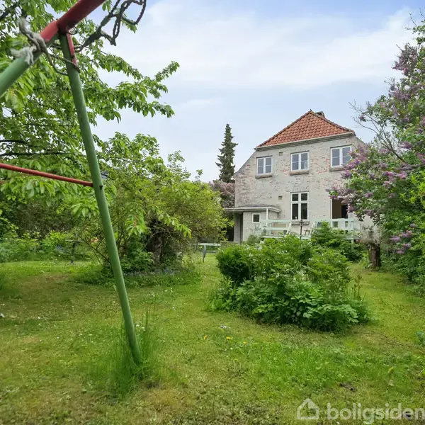 Hus står i en frodig have med græs og træer. En metalstolpe skråner i forgrunden. Klinket hus med rødt tag og store vinduer i baggrunden.
