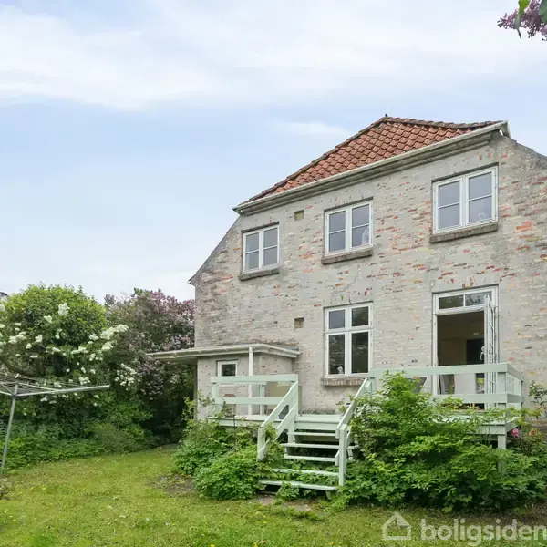 Et to-etagers hus med hvid facade og rødt tag står i en frodig have. Trapper fører op til en terrasse, omgivet af blomstrende buske.
