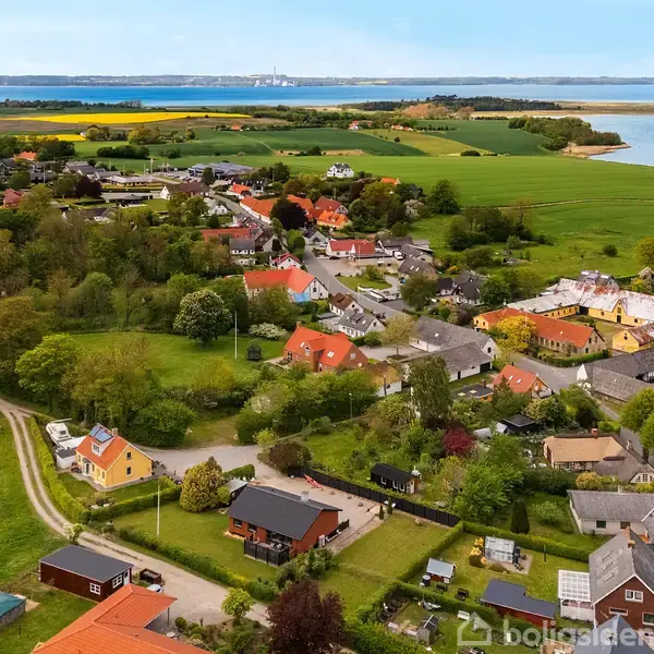 Bebyggelse med farverige huse omgivet af grønne marker og træer. Husene ligger tæt i et landsbyområde, med udsigt til havet i baggrunden. En snoet vej fører gennem landskabet.