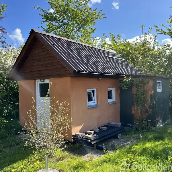 Et rundt metalbord med tre matchende stole står på en terrasse ved siden af et mørkt træhus. Haven omkring er frodig med træer og buske under en blå himmel.