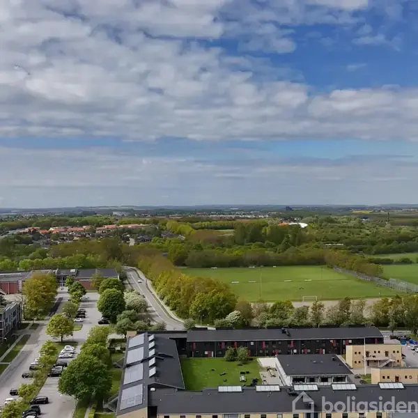 Bygninger står stille langs en vej med træer, mens en åben grøn mark og en by strækker sig i horisonten under en delvist skyet blå himmel.