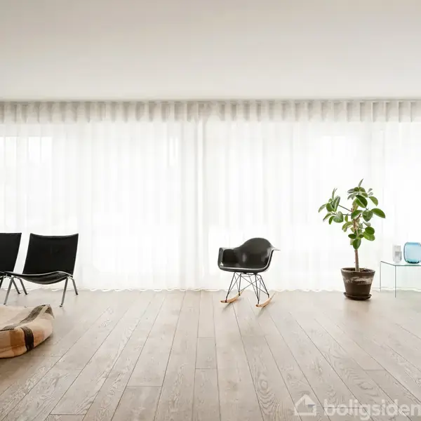Et moderne opholdsrum med trægulve, hvor to sorte stole og en gyngestol står foran store vinduer med hvide gardiner. En plante og en lænestol er også til stede.