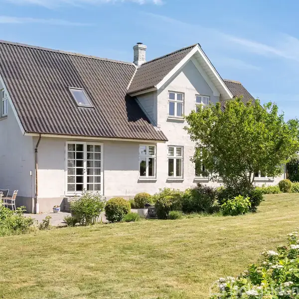 Hvid villa med sort tag står stille i en solrig have. Omringet af frodige træer og buske på en velplejet græsplæne.