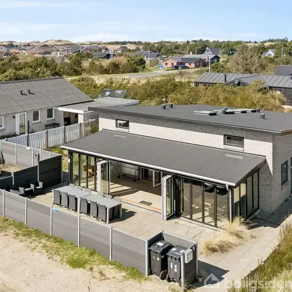 En moderne villa med fladt tag og store vinduespartier står omkranset af sand og græs i et kystnært villakvarter. Der er flere biler parkeret foran huset.
