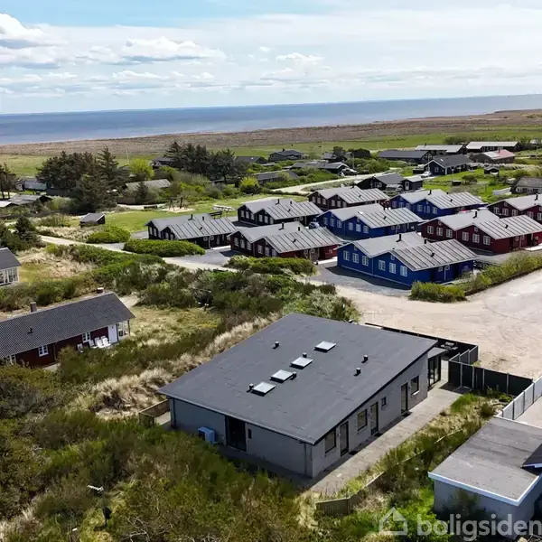 Et sommerhus med sort tag står midt i et sommerhuskvarter nær kysten. Andre huse i forskellige farver omgiver det. Baggrunden viser havet og en blå himmel.