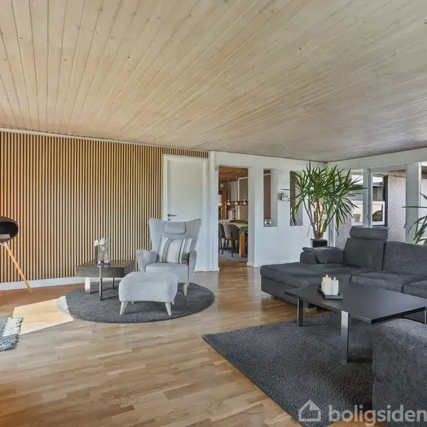 En moderne stue med en grå sofa er placeret på et mørkt tæppe. Et rundt bord og moderne lænestol står mod en væg med træpaneler. Store vinduer tillader naturligt lys.