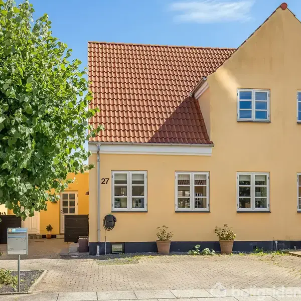 Gult hus med rødt tegltag og nummer 27 står i en boligkvarter. Foran huset er der en parkeringsplads og en træ, med klare himmel i baggrunden.