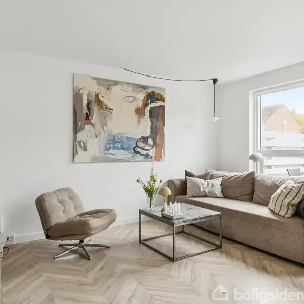 En stue med en sofa og lænestol i beige farver. Sofabordet er dekoreret med lys og en vase. Et maleri hænger på væggen, og der er hylder med dekorationer.