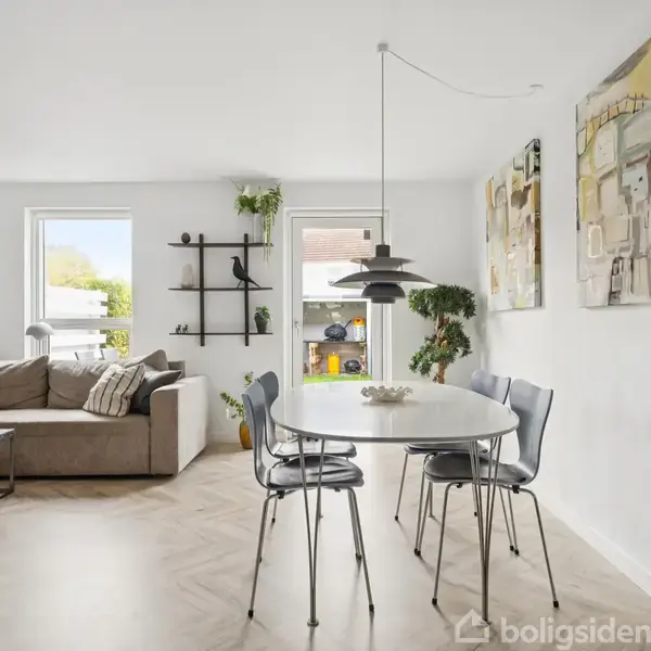 En lys stue med en beige sofa til venstre, dekoreret med puder. Til højre står et rundt spisebord med stole. Der hænger lamper og vægkunst, mens vinduerne bringer dagslys ind.