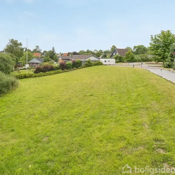 Grøn græsplæne strækker sig langs en vej med fortov til højre, omgivet af buske og træer, i et villakvarter med huse i baggrunden under en klar blå himmel.